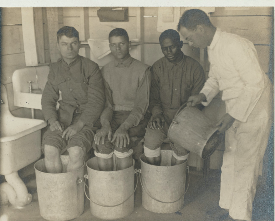 The black and white photo shows three men in uniform seated with their legs in buckets as a fourth man stands and pours water into the bucket nearest to him.