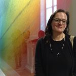 Bess Williamson, a white woman with straight brown hair, stands and smiles at the camera. She is wearing glasses. Behind her is an art exhibit of strings in the color of a rainbow. They cast a shadow on her face. 