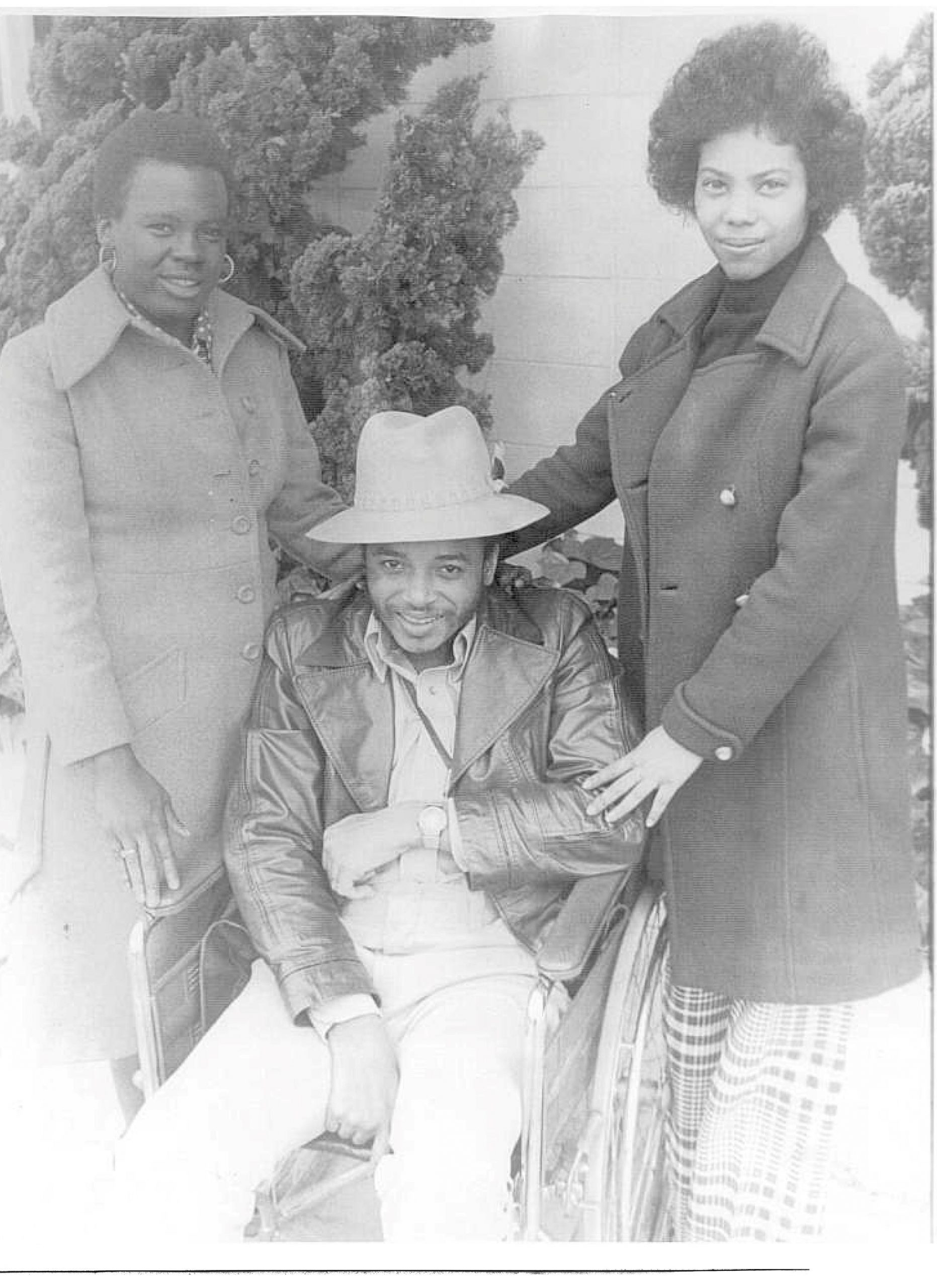 Two Black women, Barbara P. and Arlene C. standing on either side of Brad Lomax, a Black man who is seated in a wheelchair.