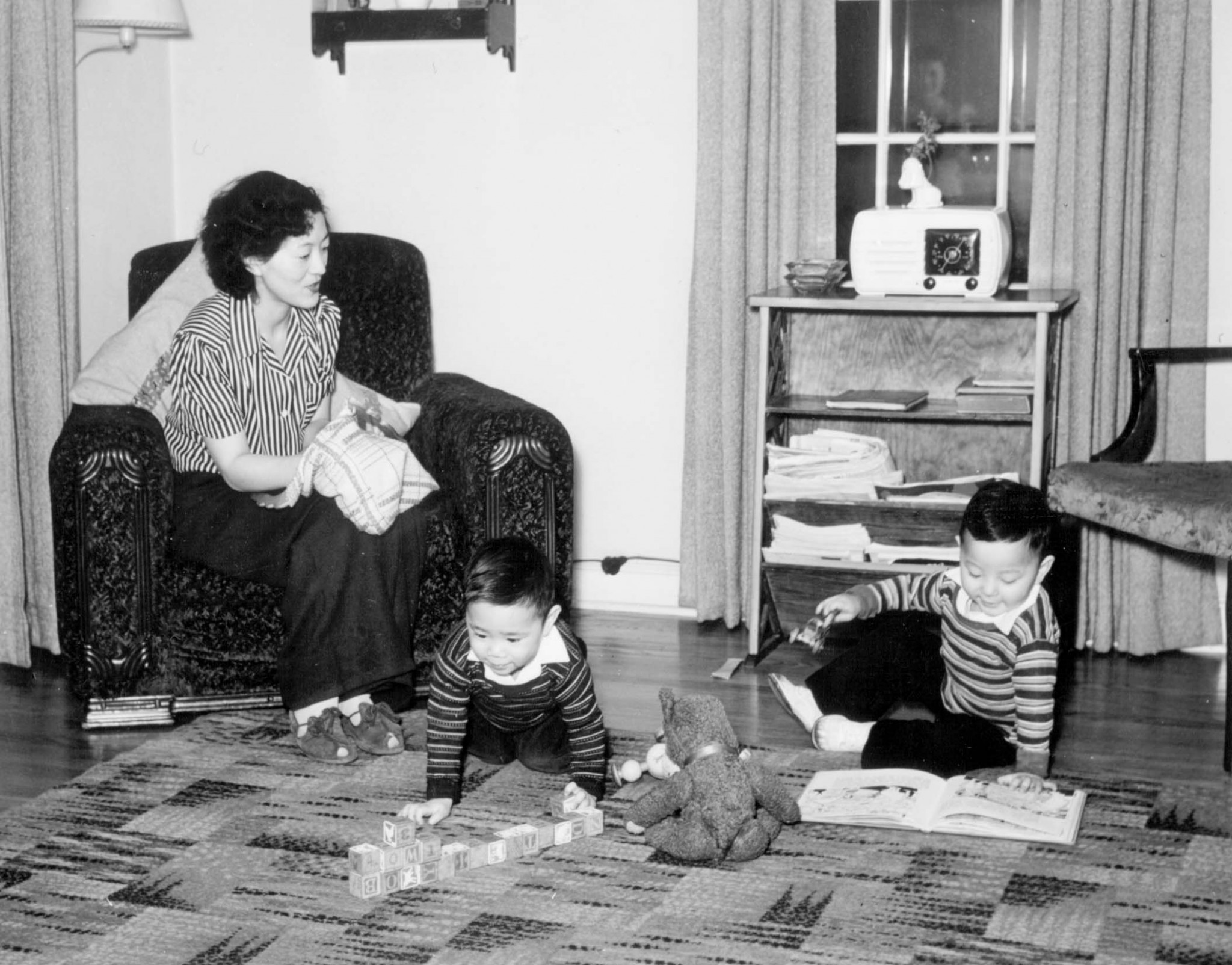 Photograph of a japanese american woman sitting on a sofa in the interior of her home, while two small boys are playing blocks and reading a book on the carpeted floor.