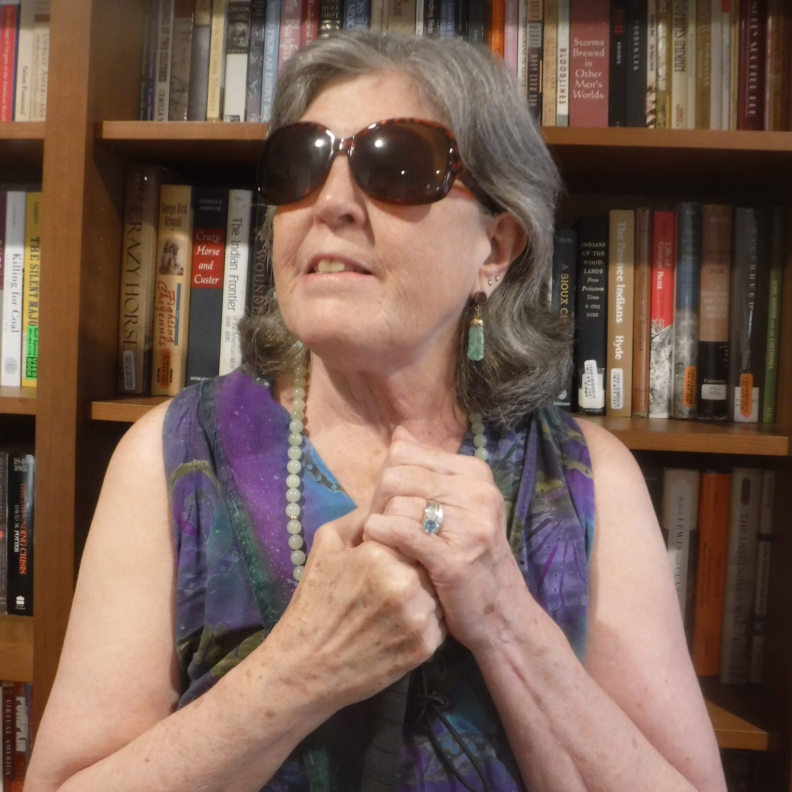 The author, a white woman with short grey hair, stands in front of a bookshelf. She is wearing dark sunglasses and holding on to a cane.