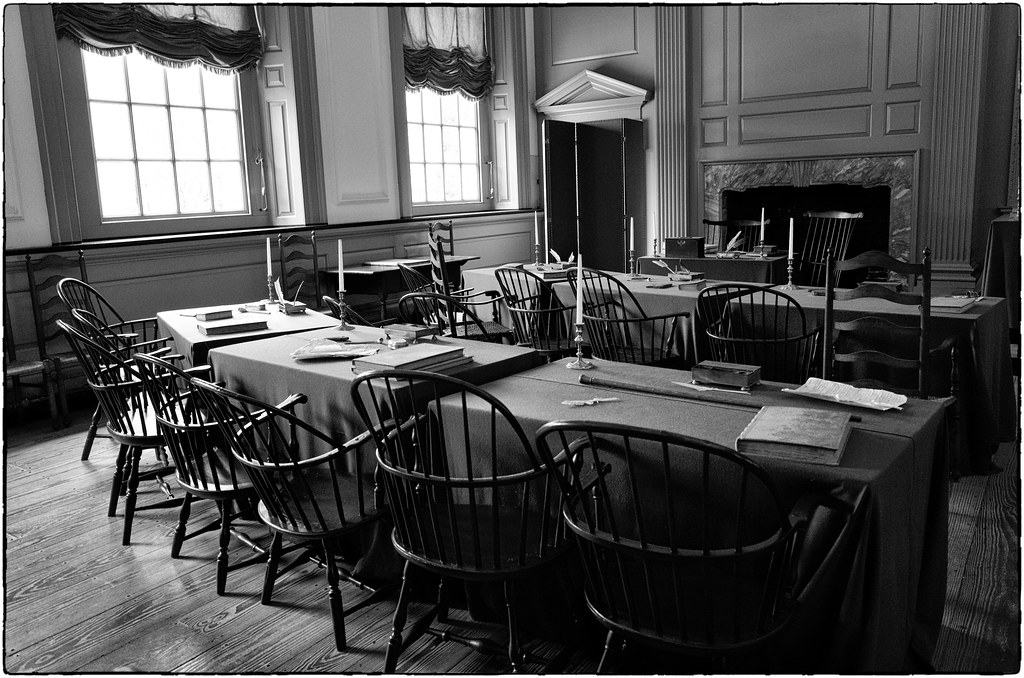 A picture of Independence Hall in Philadelphia. The picture is in black and white with a wooden floor and painted white wooden walls. The bottoms of the rectangular windows are letting sunlight through but the top is covered by fancy curtains. Numerous wooden chairs are situated at two tables with candle sticks, quills and books. A chair all the way up front is situated at a smaller table in front of the fireplace, indicating where the President of the Continental Congress sat. 