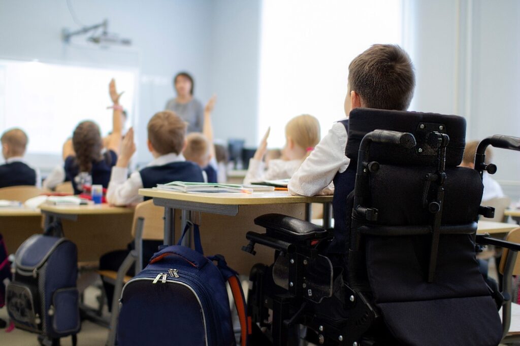 Young boy in wheelchair in classroom. Three rows of students ahead of the young boy are raising hands and looking to the teacher. The young boy in the wheelchair also looks engaged.