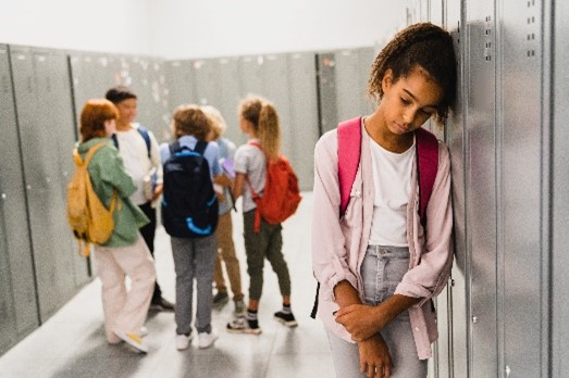 A young girl in foreground leaning against a school locker looking sad and isolated as she is excluded from a bigger group of 5 children talking with one another in the background.