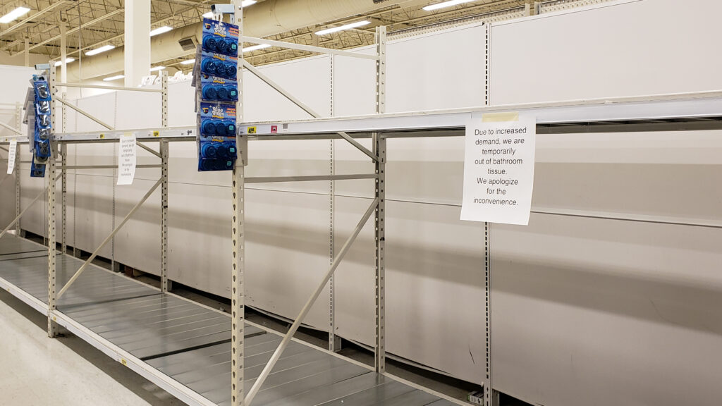 A long row of empty shelves with a flyer stating: "Due to increased demand, we are temporarily out of bathroom tissue. We apologize for the inconvenience."
