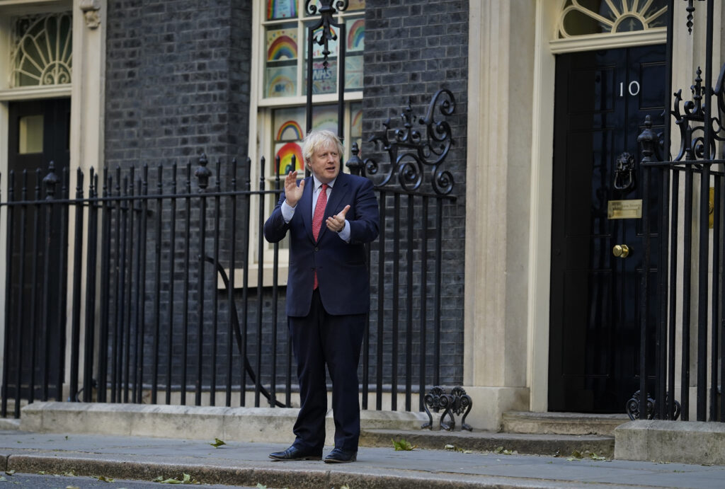 Pictured is UK Prime Minister Boris Johnson on the steps of 10 Downing Street taking part in the "Clap for our Carers." He is wearing a dark navy blue suit and a red tie.