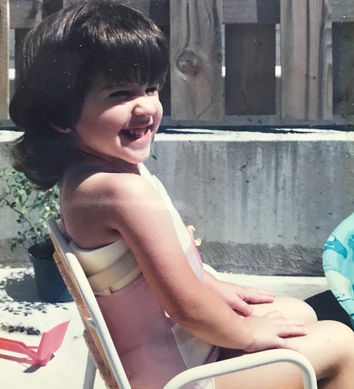 A young girl sits on a chair besides a pool. She has short dark hair and is wearing a chest brace. She is grinning.