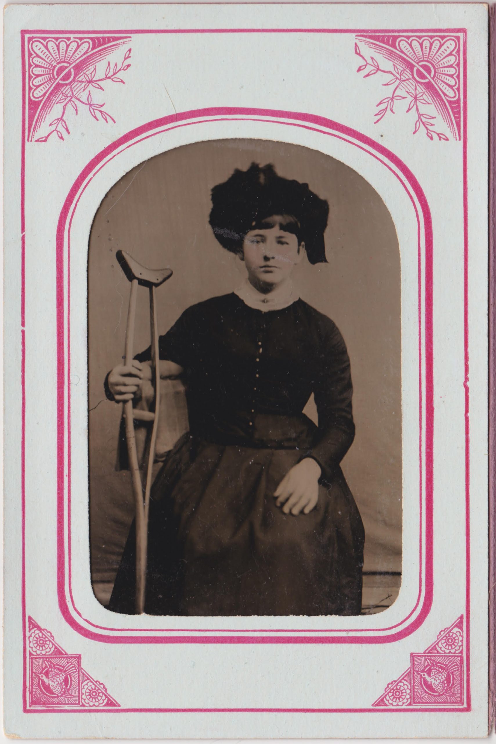 This is a black and white tintype of a woman sitting with a crutch in her right hand. Her left hand is resting on her knee. She is wearing a hat and gown with buttons (colored gold) down the bodice. The tintype is framed with white and red paper border. The border features stylized floral and bird designs in the corners.