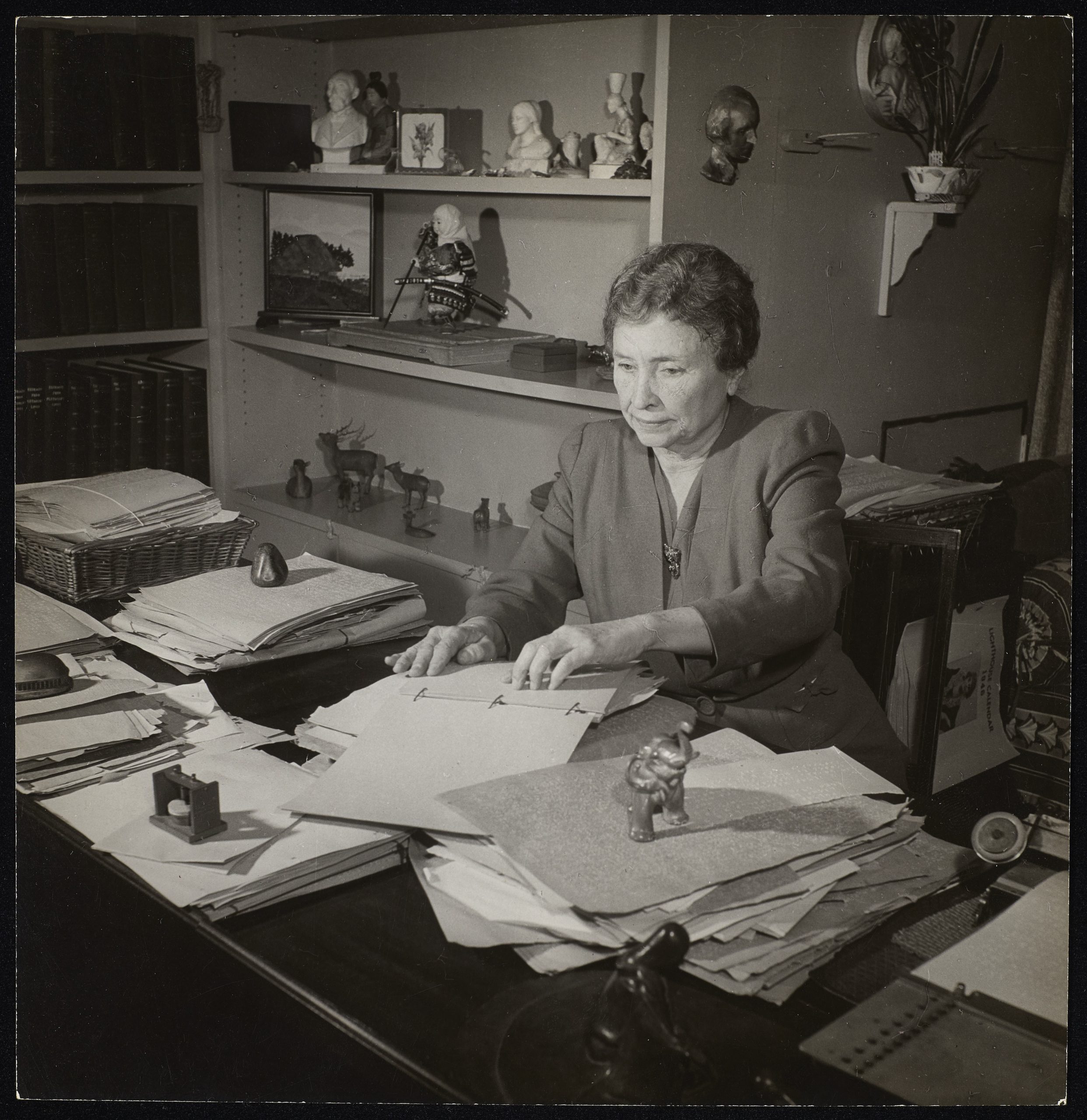 Helen Keller is reading text in braille. She is seated at her desk at her home.