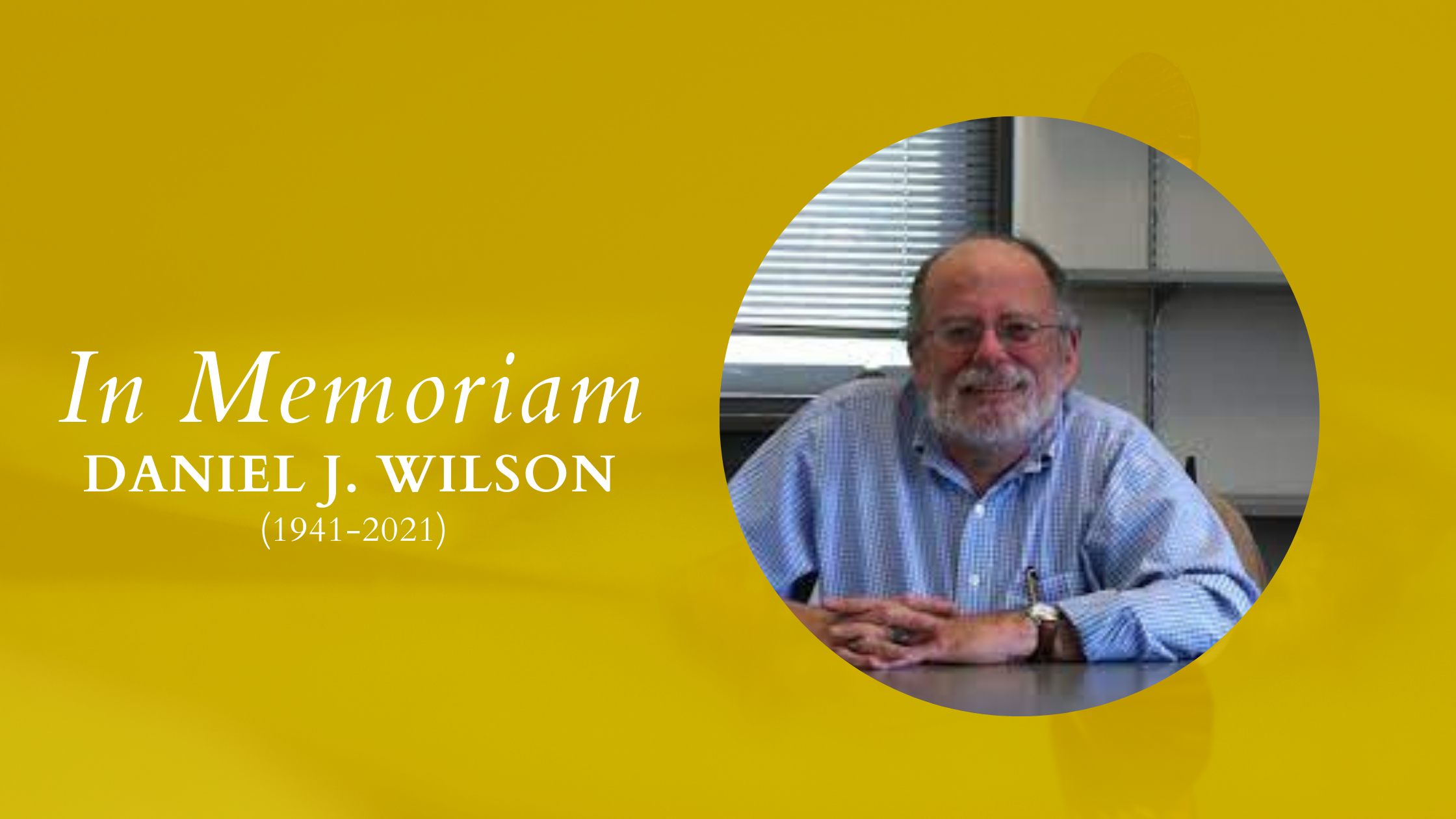 A header banner with yellow graphic on the left, upon which white text is placed: "In Memoriam Daniel J. Wilson (1941-2021)" On the right is a photograph of Dan, a bespectacled white man with white hair and beard. He is wearing a blue collared shirt, siting at a desk with his hands resting and interlocked, and smiling.