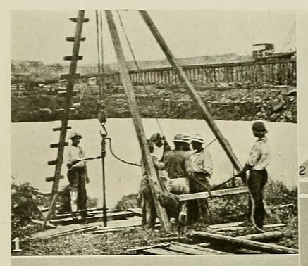Black-and-white image showing about seven Black men working under a small wooden derrick equipped with a hoist. They are wearing trousers, light shirts, and hats. One man is helping to guide a pole hanging from the hoist's lifting hook. He is wearing a white artificial leg that rises just above knee height.