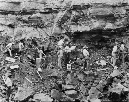 Black-and-white image showing about a dozen Black men standing next to a rocky cliff face. The men are slim, and most wear trousers, white shirts, and hats. Some of the men are holding long poles, while empty wooden dynamite boxes are scattered on the rubble around them.
